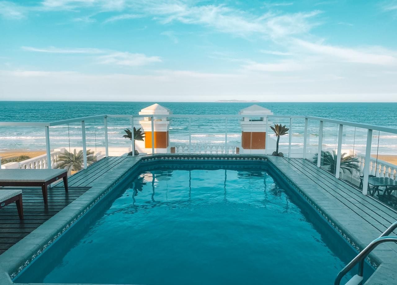 A swimming pool with a view of the ocean, surrounded by a deck with chairs and a railing.