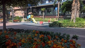 Un terrain de jeux équipé d’un toboggan et de structures d’escalade, entouré de jardins aménagés et de palmiers.