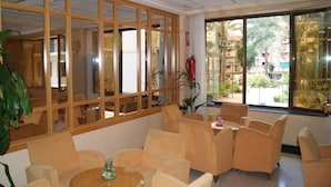 Un hall d’hôtel avec des fauteuils beiges disposés autour d’une table centrale, de grandes fenêtres donnant sur l’extérieur et un extincteur fixé au mur.