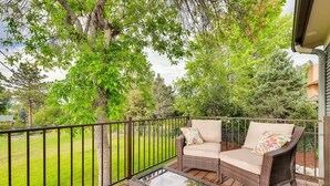 A covered outdoor seating area with wicker furniture and a glass table, surrounded by a metal railing and a view of a grassy yard and trees. - Discovery Park House (Wheat Ridge)