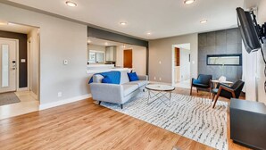 A modern living room with a sofa, coffee table, and chairs. There is a kitchen area visible through an open doorway. - Discovery Park House (Wheat Ridge)