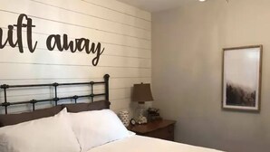A neatly made bed with white linens, a headboard with horizontal lines, a bedside table with a lamp, and a wall with a 'shift away' sign.