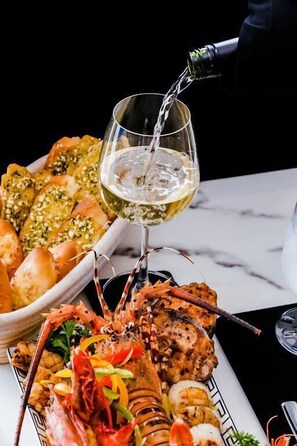 A lobster tail, grilled vegetables, and bread on a plate with a glass of white wine being poured. - Regal Réseau Hotel & Spa (Negombo)