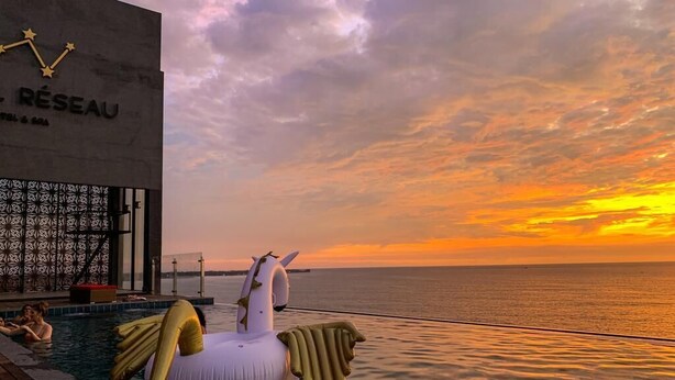 Une scène au bord de la piscine avec une grande bouée en forme de licorne gonflable, un bâtiment à l’architecture ornée d’une étoile et une vue sur le coucher du soleil.