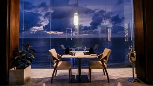 Un espace repas comprenant une table et des chaises, un luminaire suspendu et une vue sur la mer grâce à de grandes fenêtres.