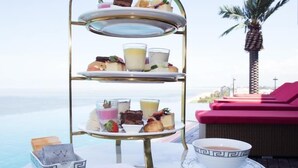 Un présentoir à trois niveaux, garni de diverses pâtisseries et boissons, un bol de thé et une serviette sur une table ronde placée au bord d’une piscine, avec un palmier en arrière-plan.