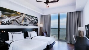 Une chambre d’hôtel avec deux lits, une grande fenêtre donnant sur la mer, un ventilateur de plafond et un bureau avec une lampe.