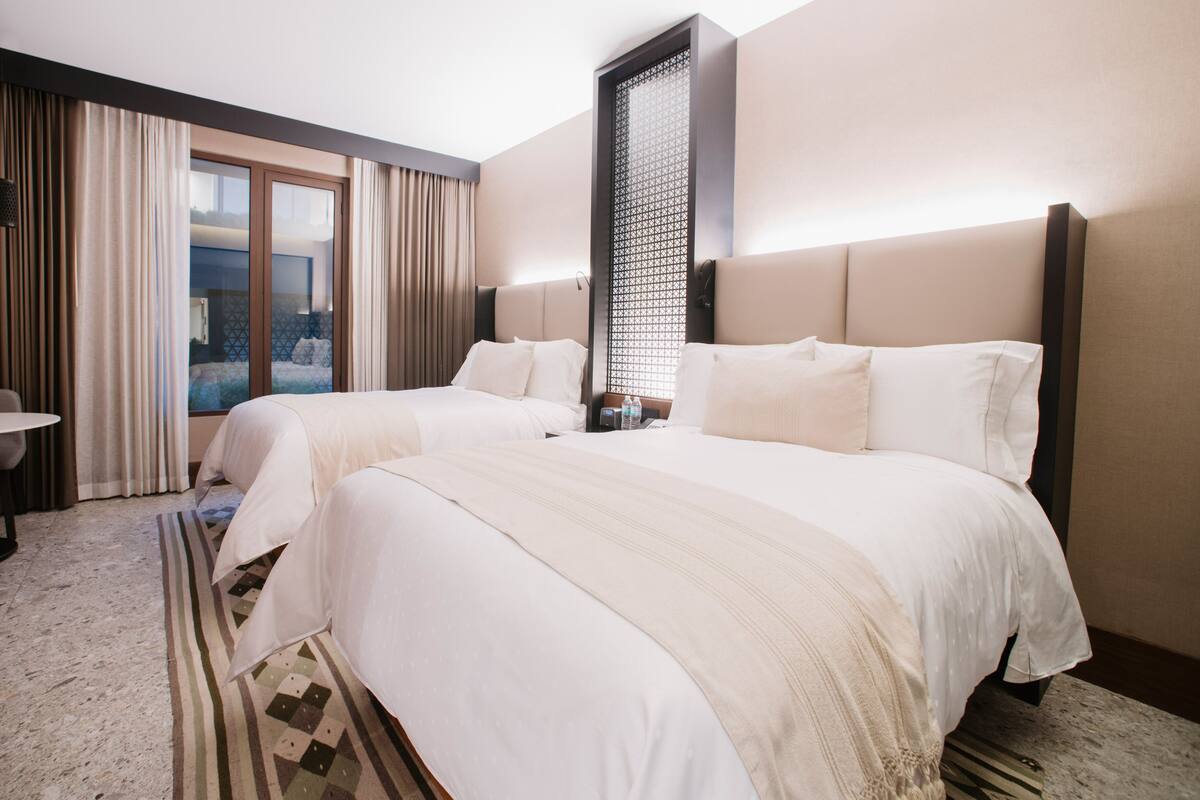 Une chambre d’hôtel avec deux lits, une grande fenêtre avec des rideaux, une vue sur la ville et une petite table.