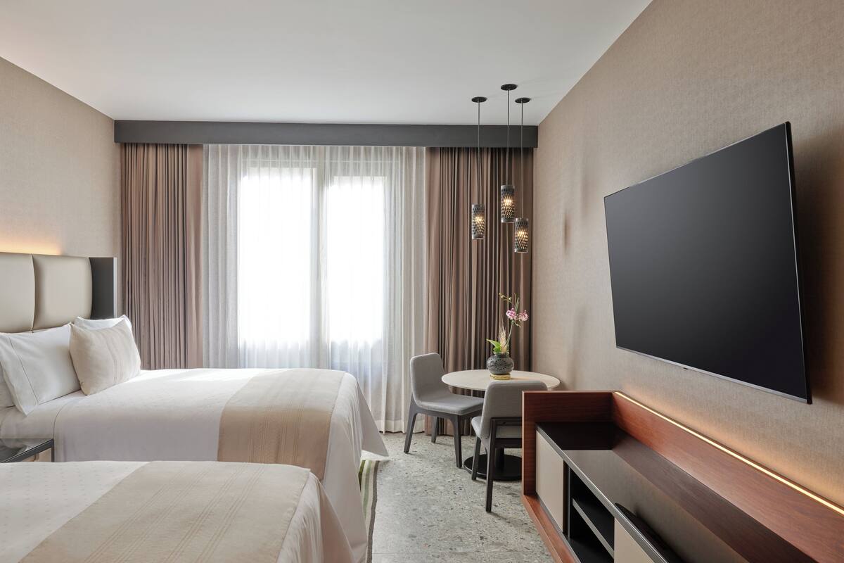 Une chambre d’hôtel avec deux lits, une télévision, un bureau avec une chaise et une fenêtre avec des rideaux.