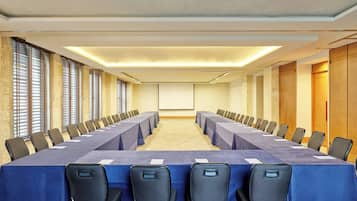 Una sala de conferencias con una larga mesa, sillas, una pantalla de proyector y grandes ventanales con persianas.