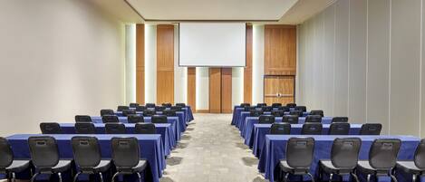 A conference room with rows of tables and chairs, a projector screen, and a large wooden door.