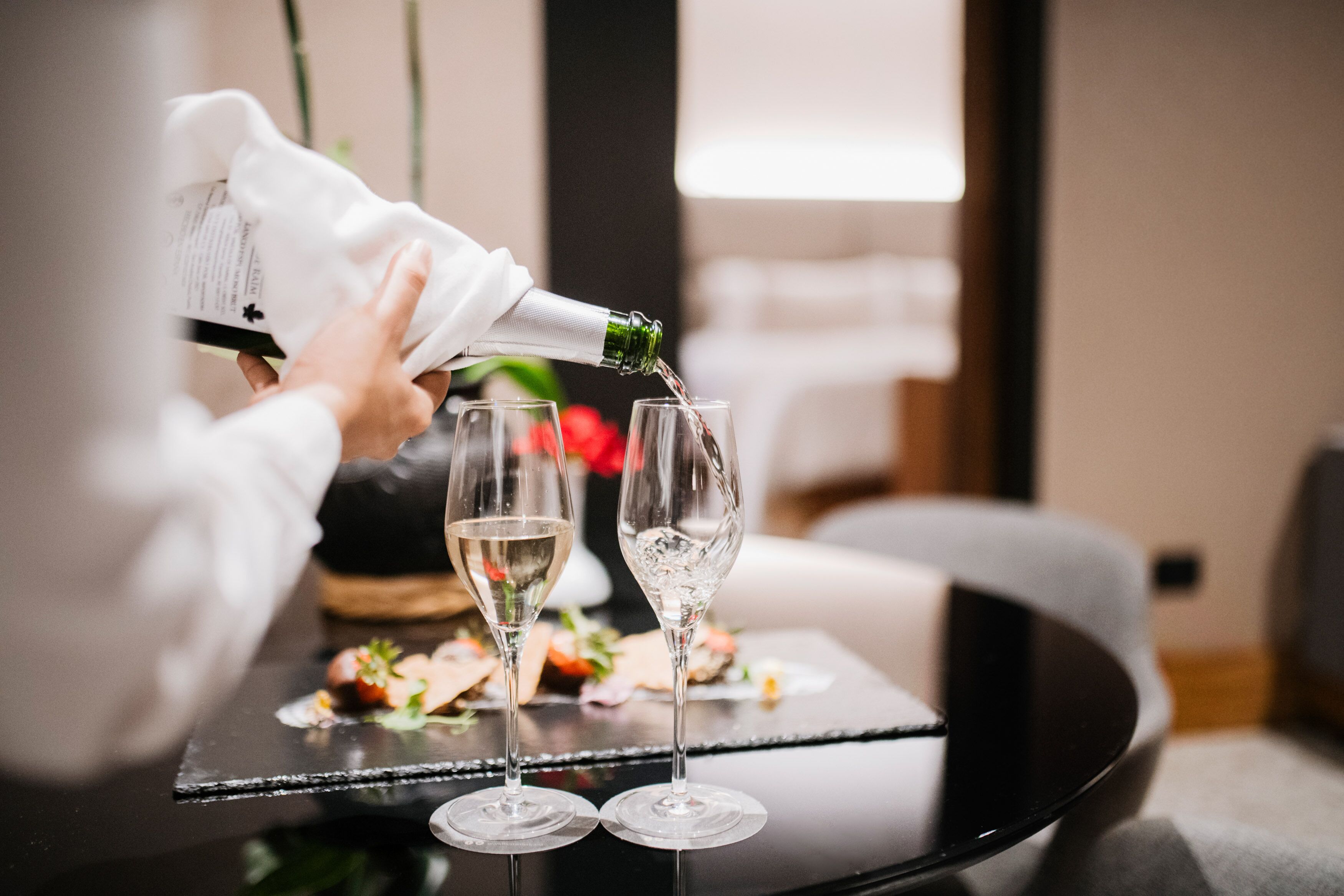 En person hælder drik i to glas på et bord, med en desserttallerken i baggrunden.