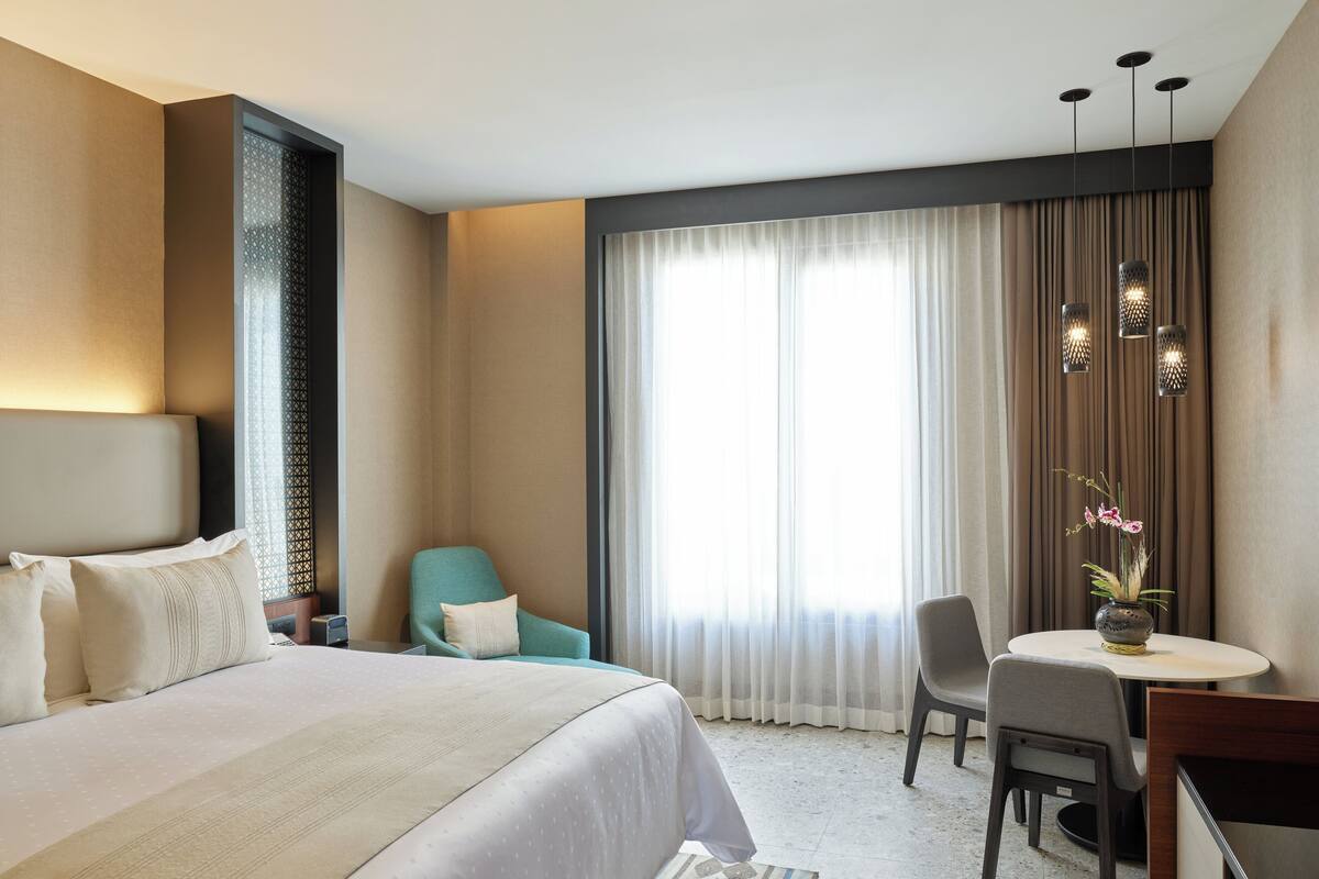 Une chambre d’hôtel avec un grand lit, un bureau avec une chaise, une petite table avec un vase de fleurs et une fenêtre avec des rideaux.