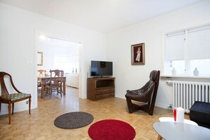 Una sala de estar con televisor, un tapete rojo y uno gris, un sillón de cuero marrón, y una zona de comedor con mobiliario de madera.