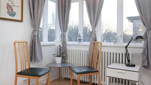 Una habitación con dos sillas de madera, una mesita pequeña, un escritorio blanco, un radiador y una ventana con cortinas.