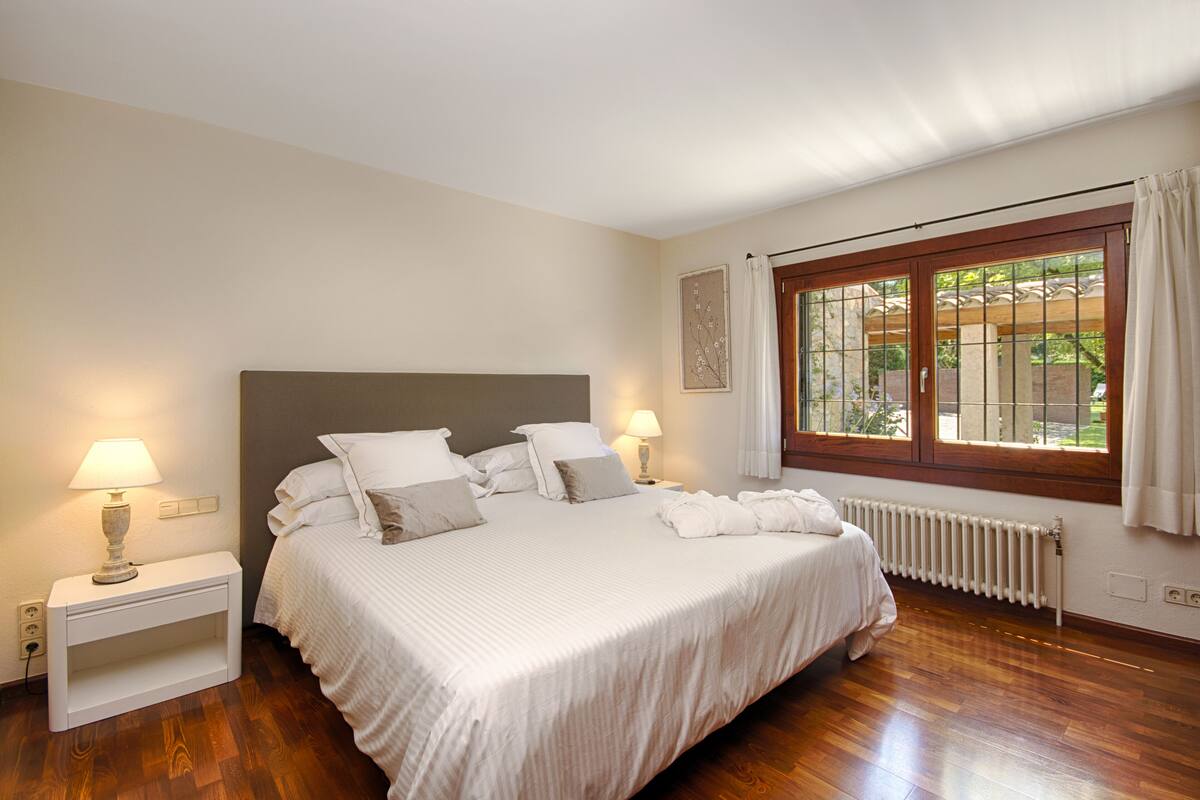 Una cama bien hecha con sábanas y almohadas blancas, una mesita de noche con una lámpara, una ventana con cortinas y un radiador.