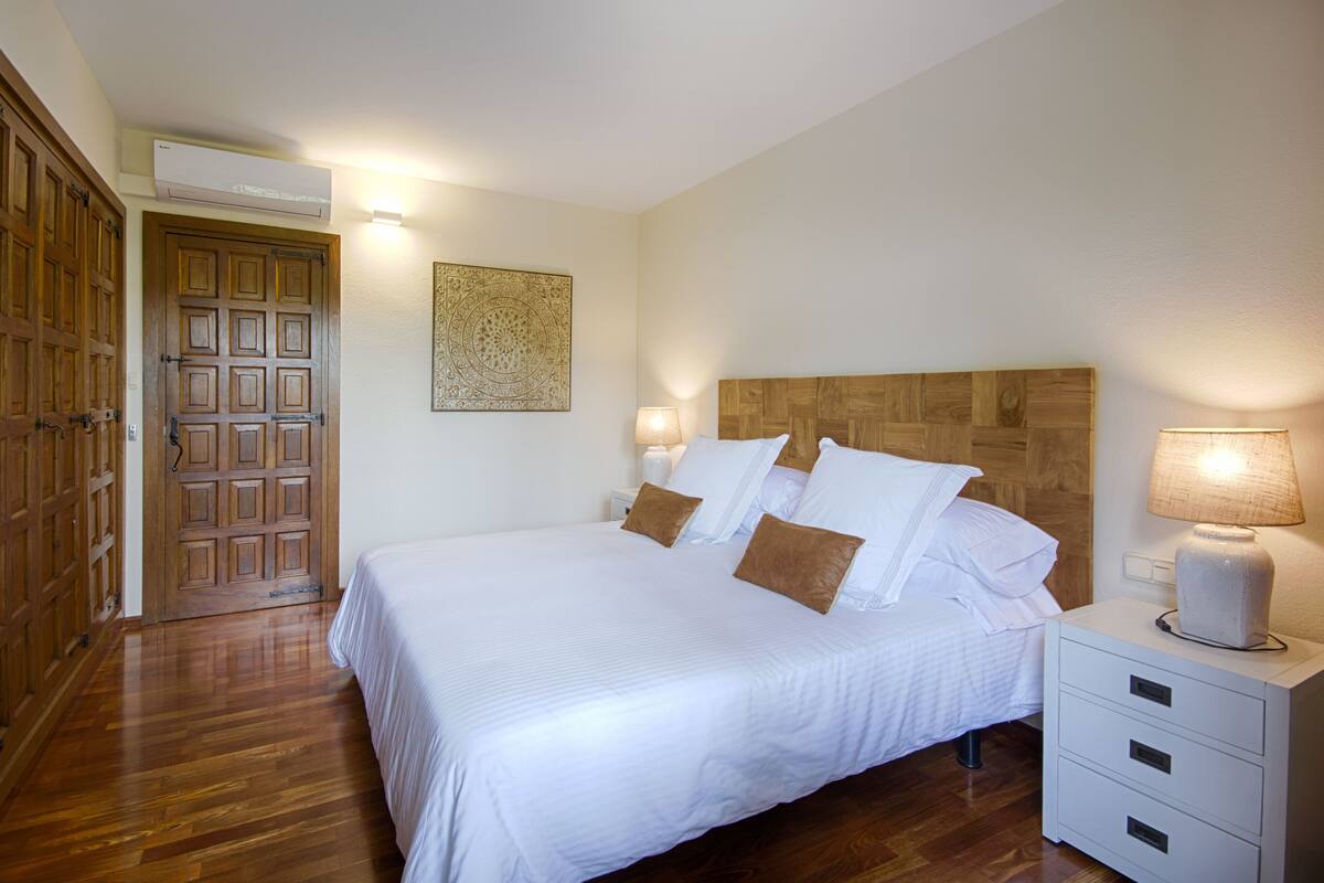 Un dormitorio ordenado con una cama grande, cabecera de madera, mesita de noche con lámpara y una puerta con panel decorativo.