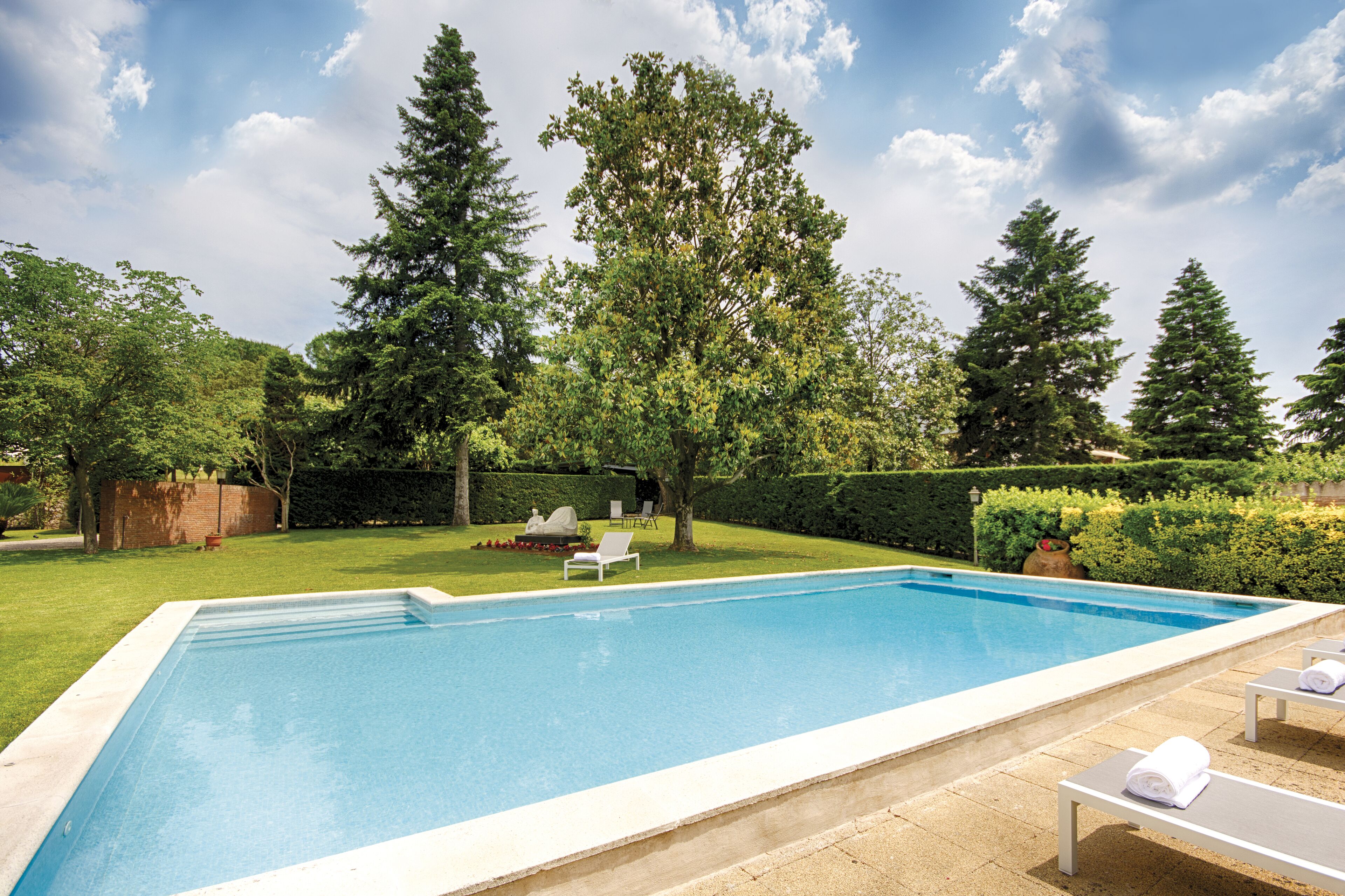 Una piscina rectangular con borde blanco, rodeada por un césped cuidado y altos árboles. Hay sillas de descanso y una pequeña mesa con una toalla.