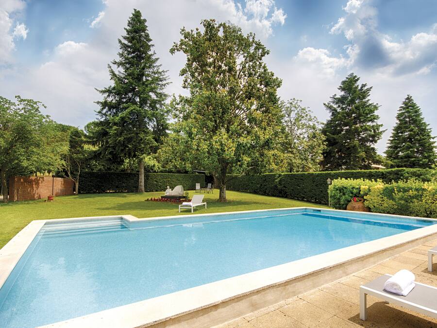 Una piscina rectangular con borde blanco, rodeada por un césped cuidado y altos árboles. Hay sillas de descanso y una pequeña mesa con una toalla.