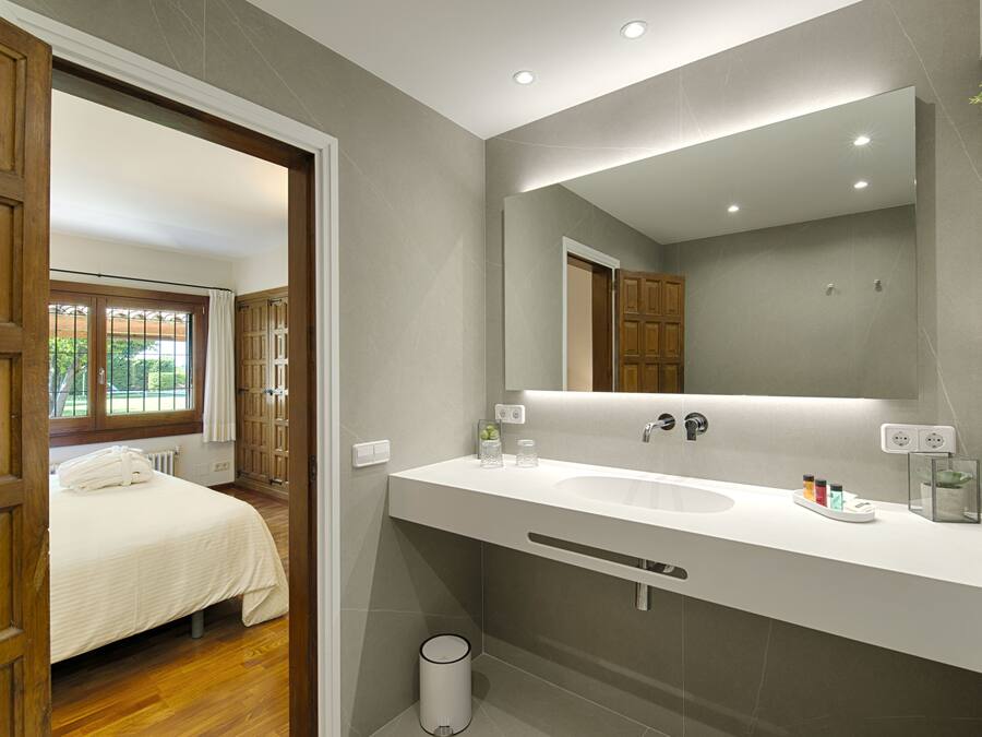 Un baño moderno con un espejo grande, un lavabo blanco y una puerta de madera que da a un dormitorio.