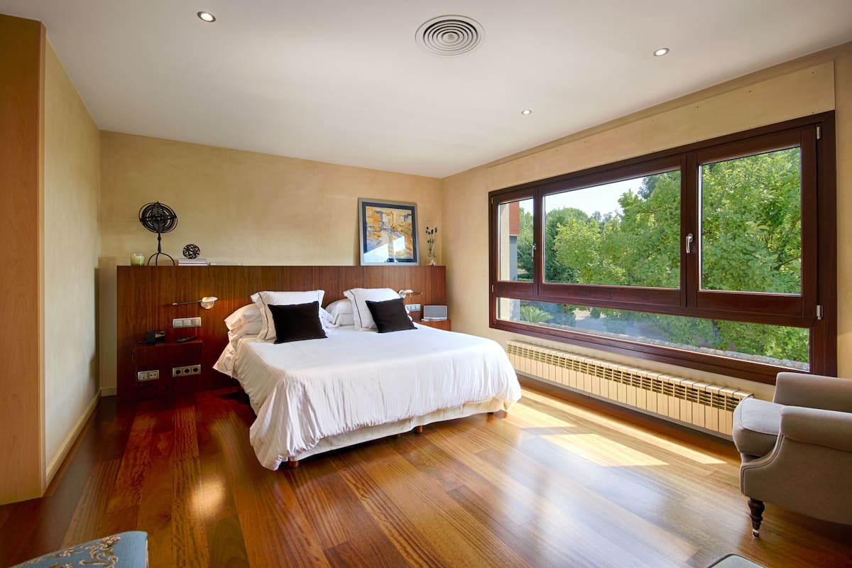 Un dormitorio con una cama grande, suelo de madera, una mesita de noche, una silla y un amplio ventanal con vistas a árboles.