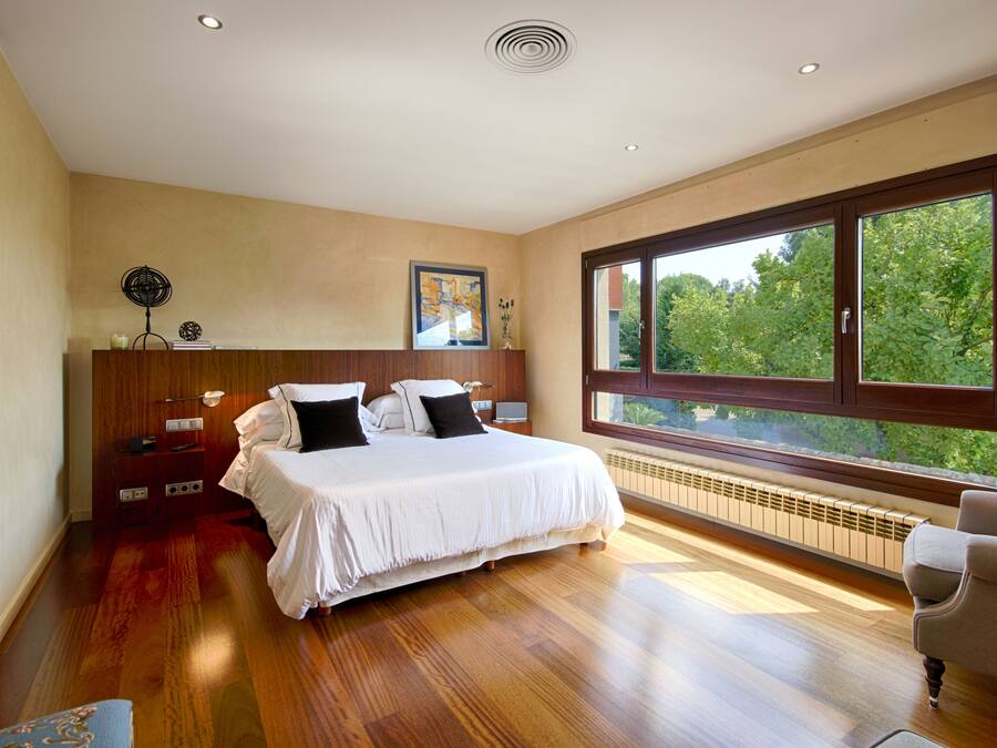 Un dormitorio con una cama grande, suelo de madera, una mesita de noche, una silla y un amplio ventanal con vistas a árboles.