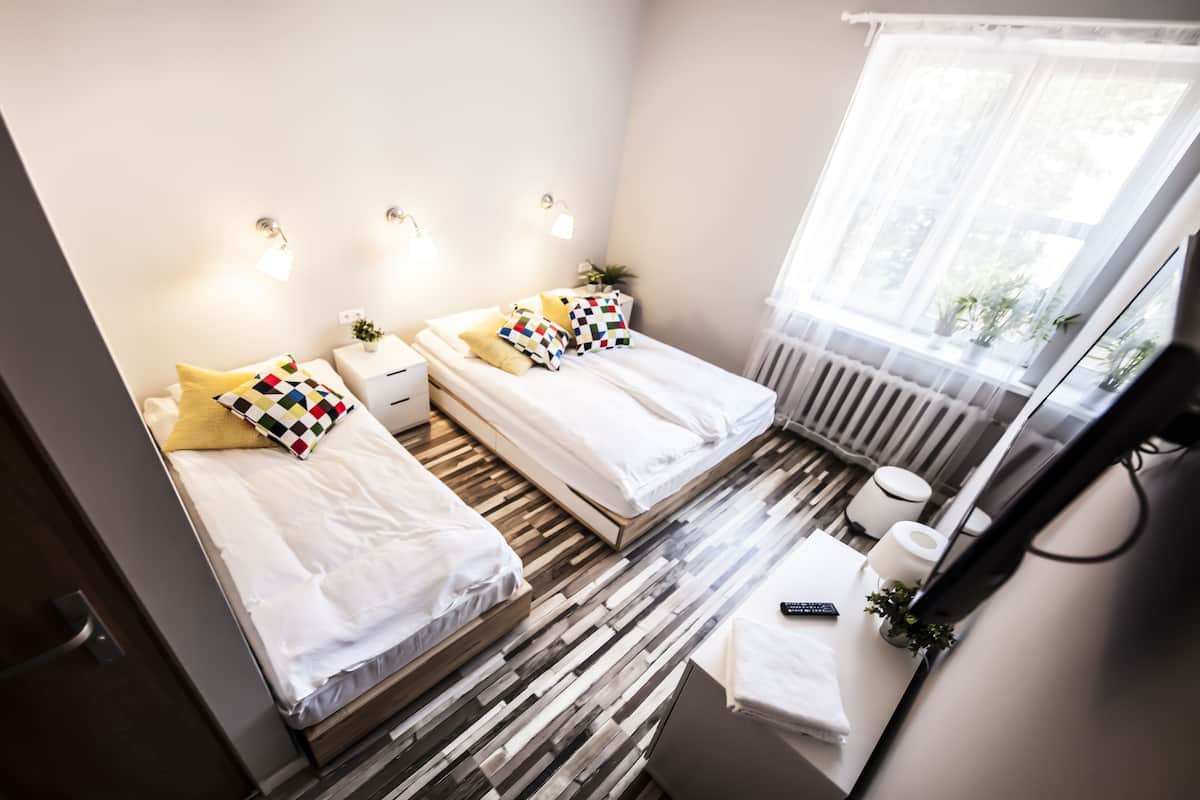 A modern hotel room with two beds, a wooden floor, a large window with a view, a bedside table with a lamp, and a mirror.