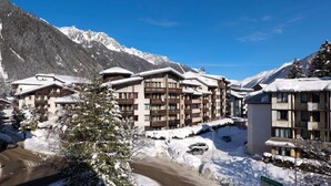Un complejo turístico en la montaña cubierto de nieve, con edificios de varios pisos, un camino sinuoso y un cielo azul despejado.
