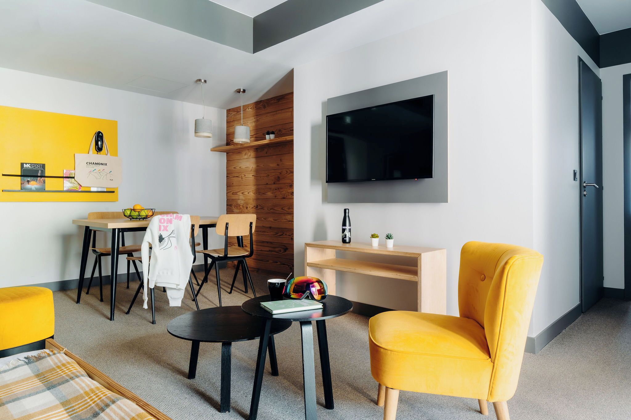A modern living room with a flat-screen TV, a dining area with wooden chairs, a yellow armchair, and a small table with a book and sunglasses.