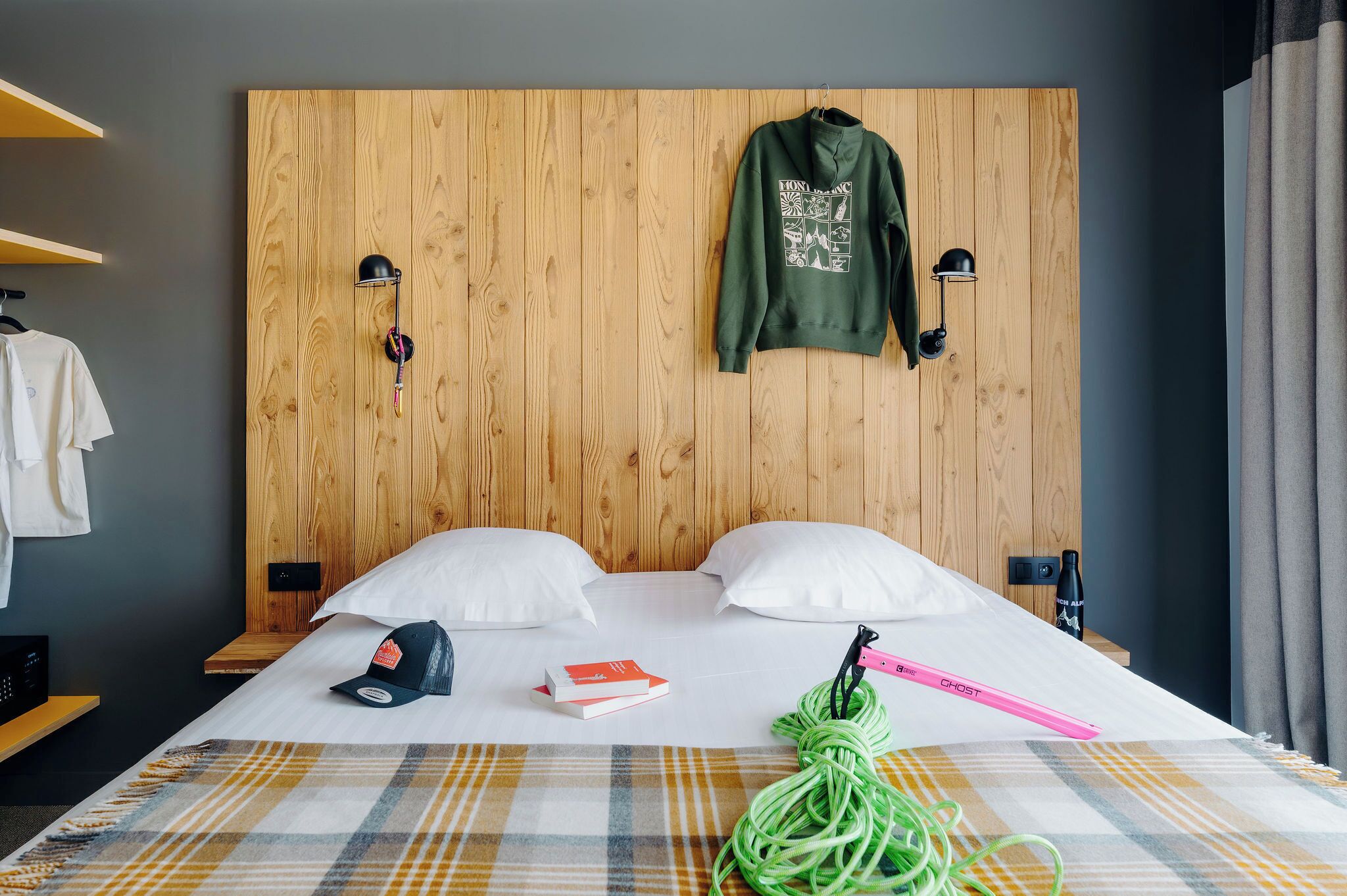 A hotel room with a wooden headboard, two pillows, a bed with a plaid blanket, a green jacket hanging on the headboard, a cap, a book, and a green cord.