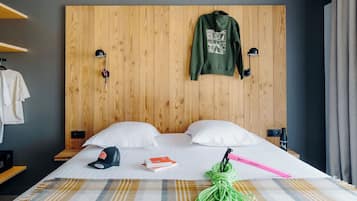 A hotel room with a wooden headboard, two pillows, a bed with a plaid blanket, a green jacket hanging on the headboard, a cap, a book, and a green cord.