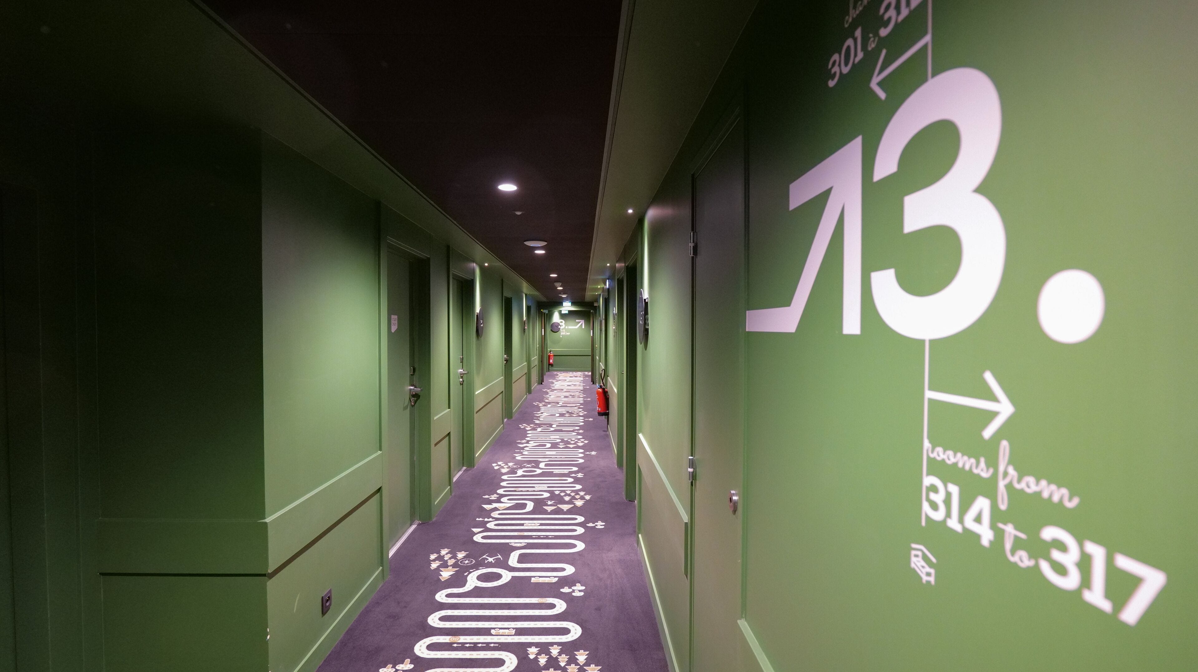 A hotel corridor with green walls, numbered room signs, and a purple carpet with white directional arrows.