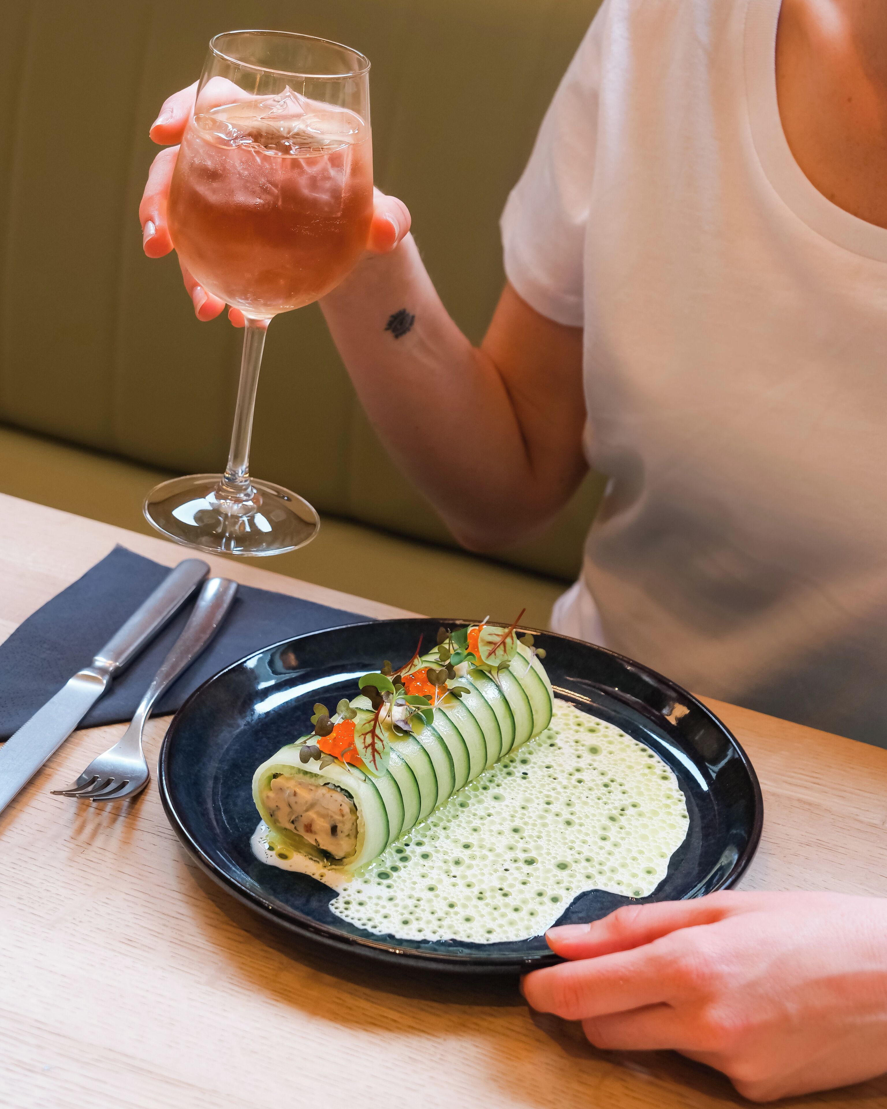 Una persona sosteniendo una copa de vino, sentada a una mesa con un plato de comida y cubiertos.