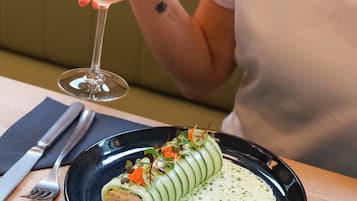 Una persona sosteniendo una copa de vino, sentada a una mesa con un plato de comida y cubiertos.