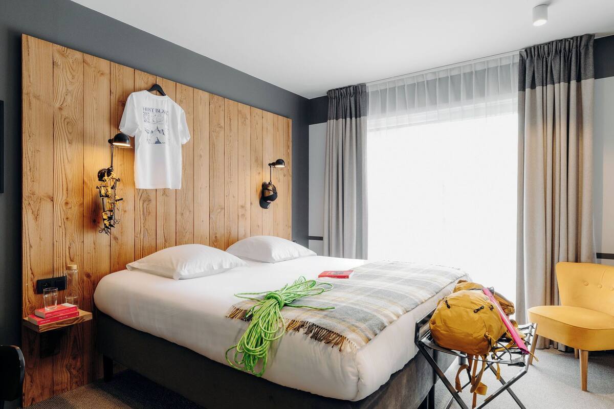 A modern hotel room with a wooden headboard, a bed with white linens, a yellow chair, and a large window with curtains.