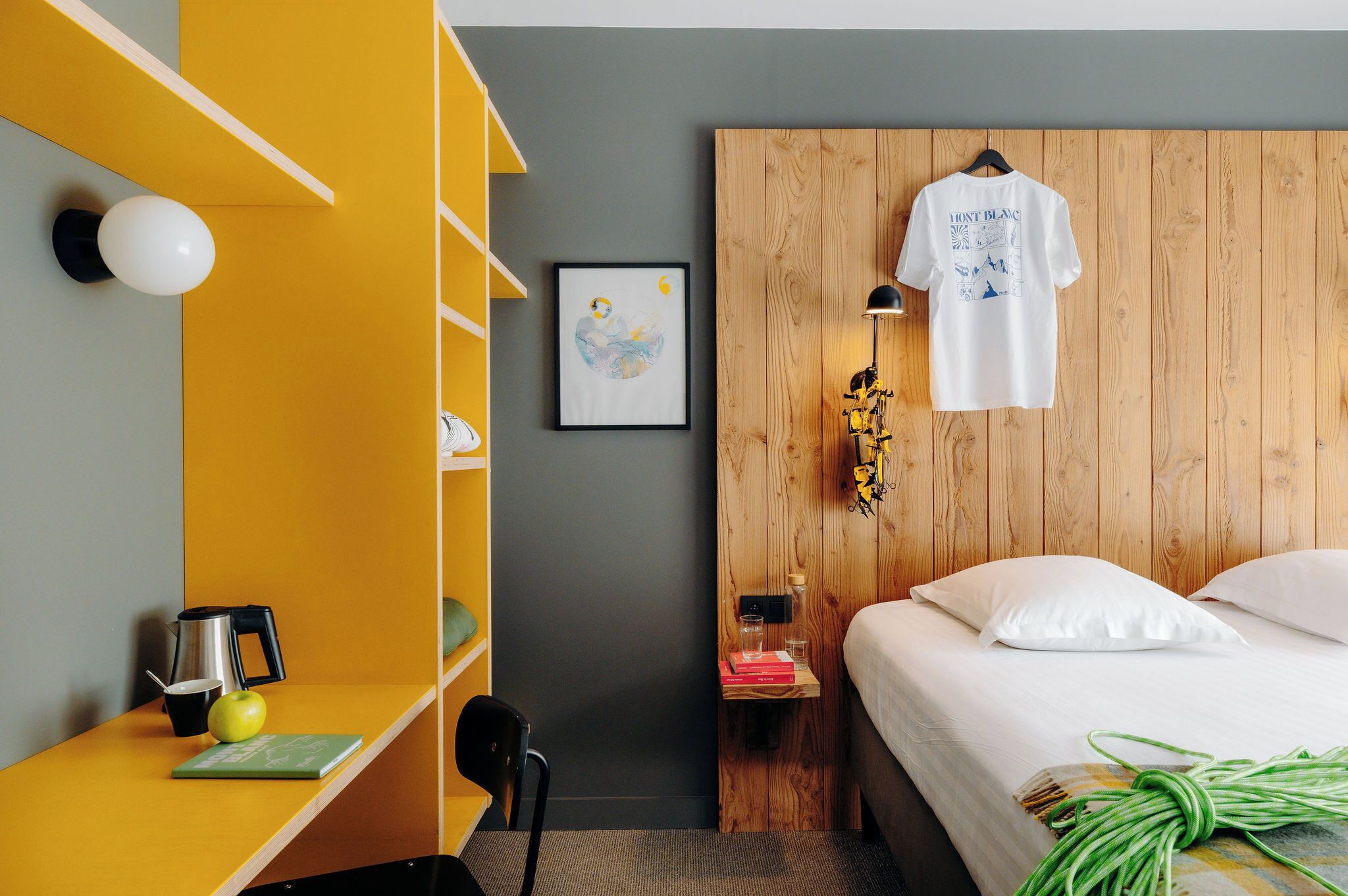 A hotel room with a bed, a wooden headboard, a yellow shelf, a framed picture, and a hanging t-shirt.