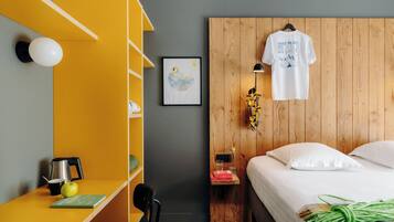 A hotel room with a bed, a wooden headboard, a yellow shelf, a framed picture, and a hanging t-shirt.