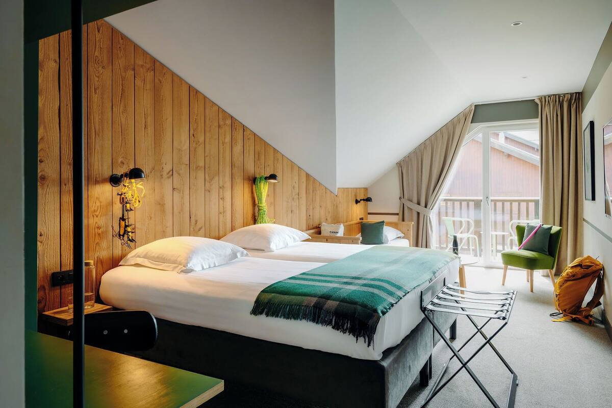 A hotel room with a wooden sloped ceiling, a bed with white linens, a green and black blanket, a small table, a chair, and a balcony with a view.