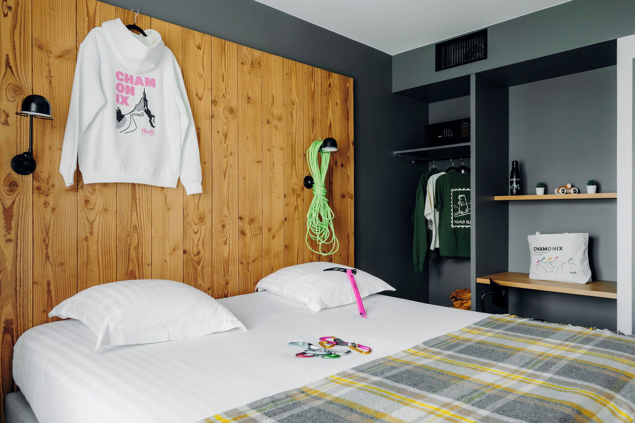 Camera d'albergo con testiera in legno, letto con biancheria bianca, coperta a quadri, asciugamano verde appeso e una borsa bianca con la scritta 'CHAMONIX'.