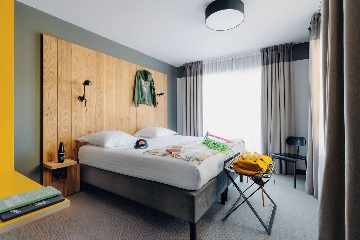 A modern hotel room with a wooden headboard, a bed with white linens, a small bedside table with a water bottle, a chair, and a desk with a lamp.