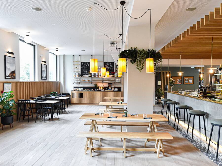 A modern restaurant with wooden tables and benches, a bar area with stools, and a kitchen in the background.