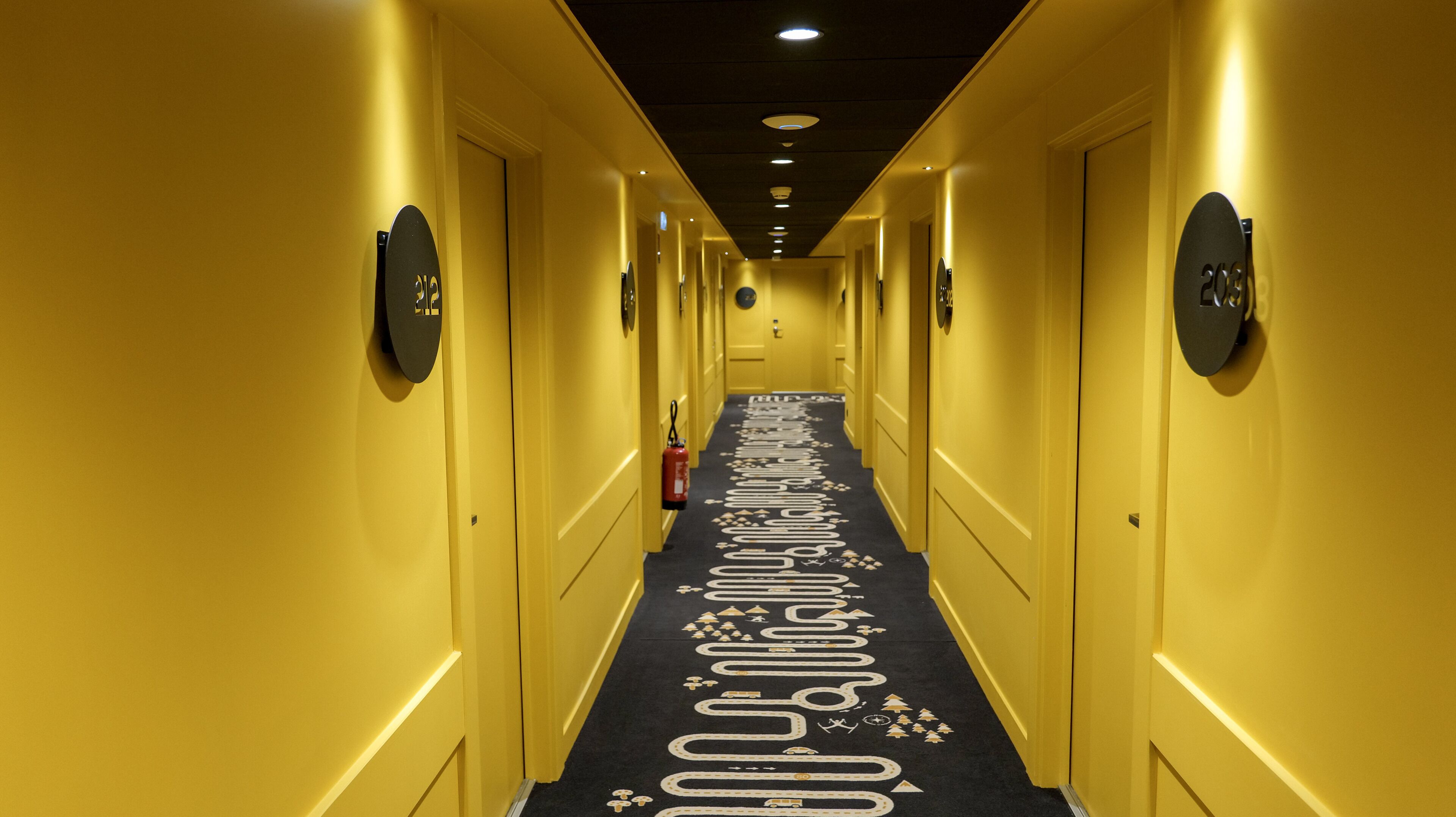 Un couloir d’hôtel avec un tapis orné d’un motif représentant une route et des véhicules, des portes portant des numéros et un extincteur fixé au mur.