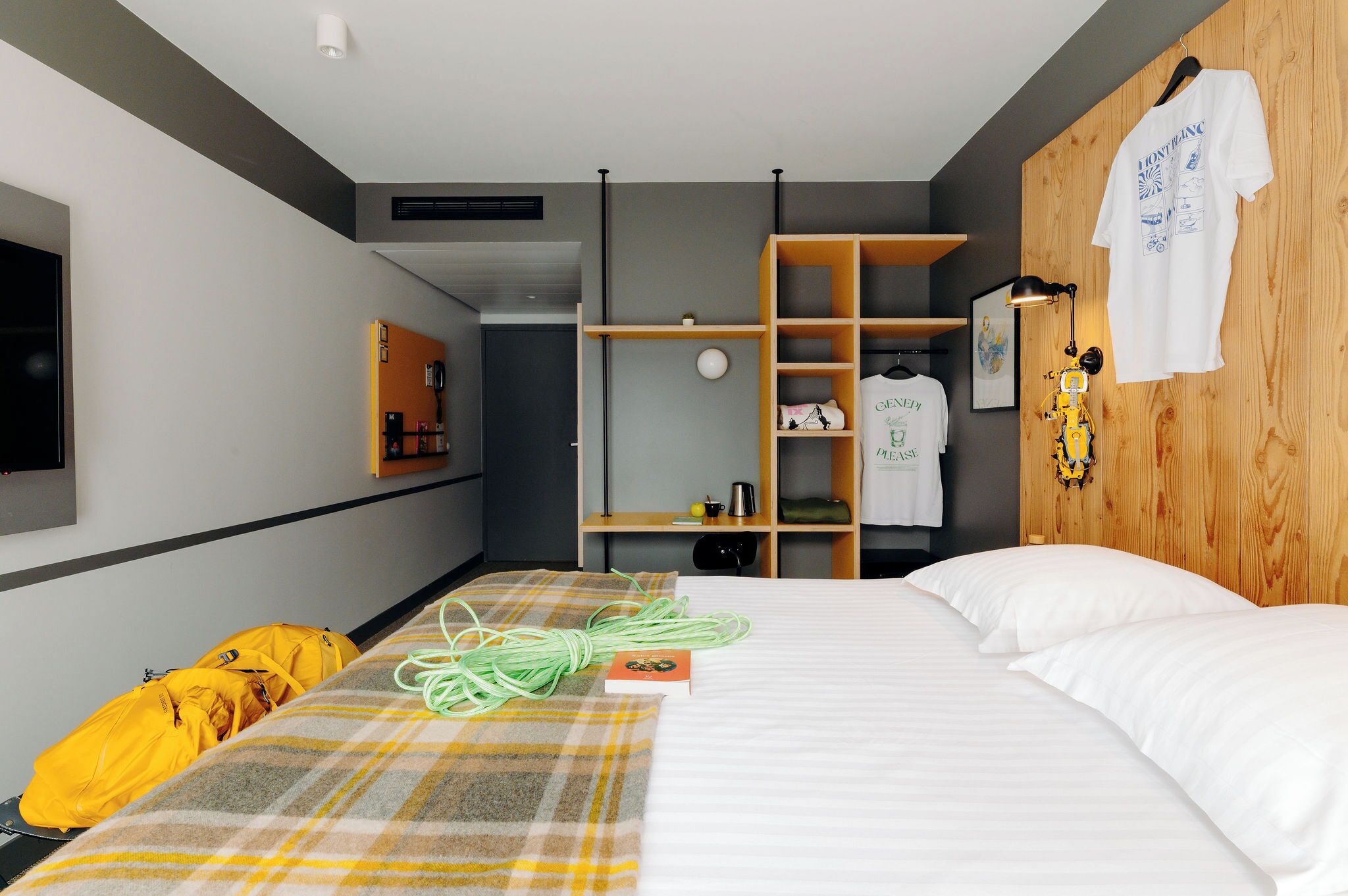 A hotel room with a bed, a wooden wardrobe, a shelf with a lamp, and a TV mounted on the wall.