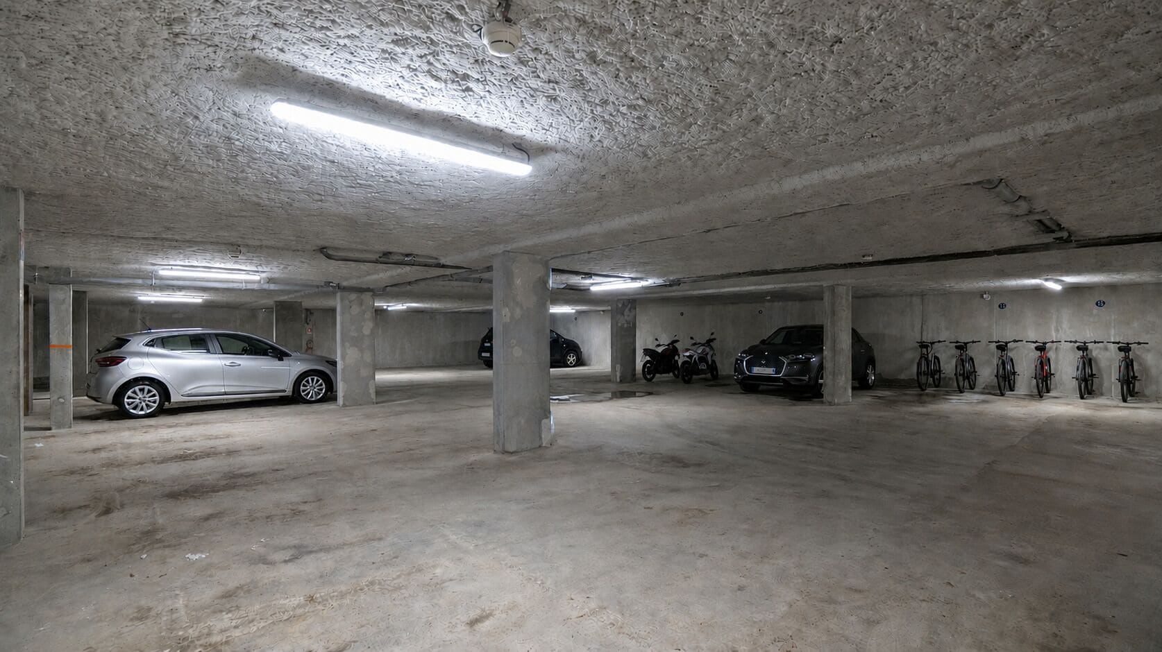 Eine mehrgeschossige Tiefgarage mit Betonpfeilern, Neonbeleuchtung und verschiedenen Fahrzeugen, darunter Autos und Fahrräder.