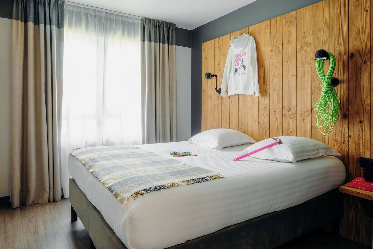 A neatly made bed with a striped blanket, a white pillow, and a pink object on it. There is a wooden wall panel with a white shirt hanging on it and a green object on the wall.