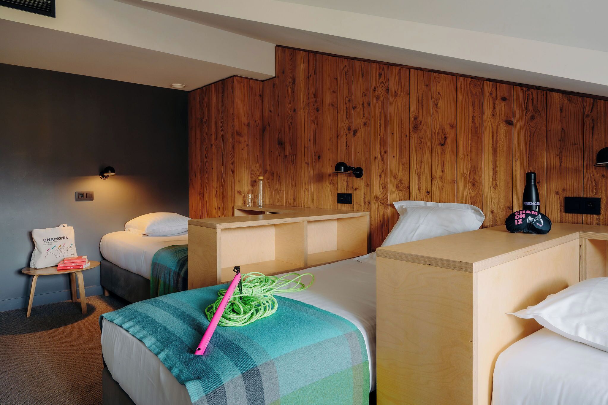 A hotel room with two beds, a wooden headboard, a small table with a bag and books, and a bottle on the nightstand.