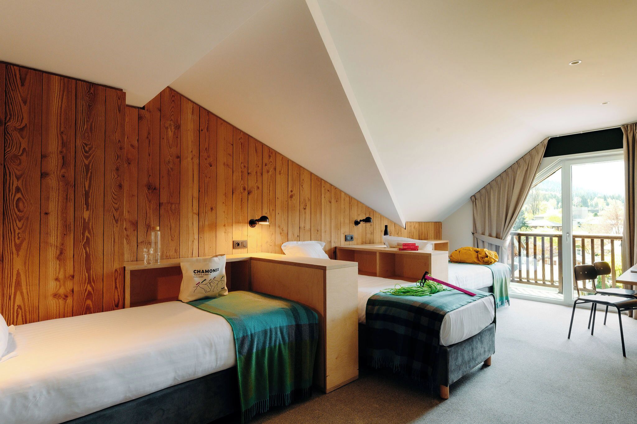 A room with two beds, a wooden wall, a balcony with a view, and a small table.