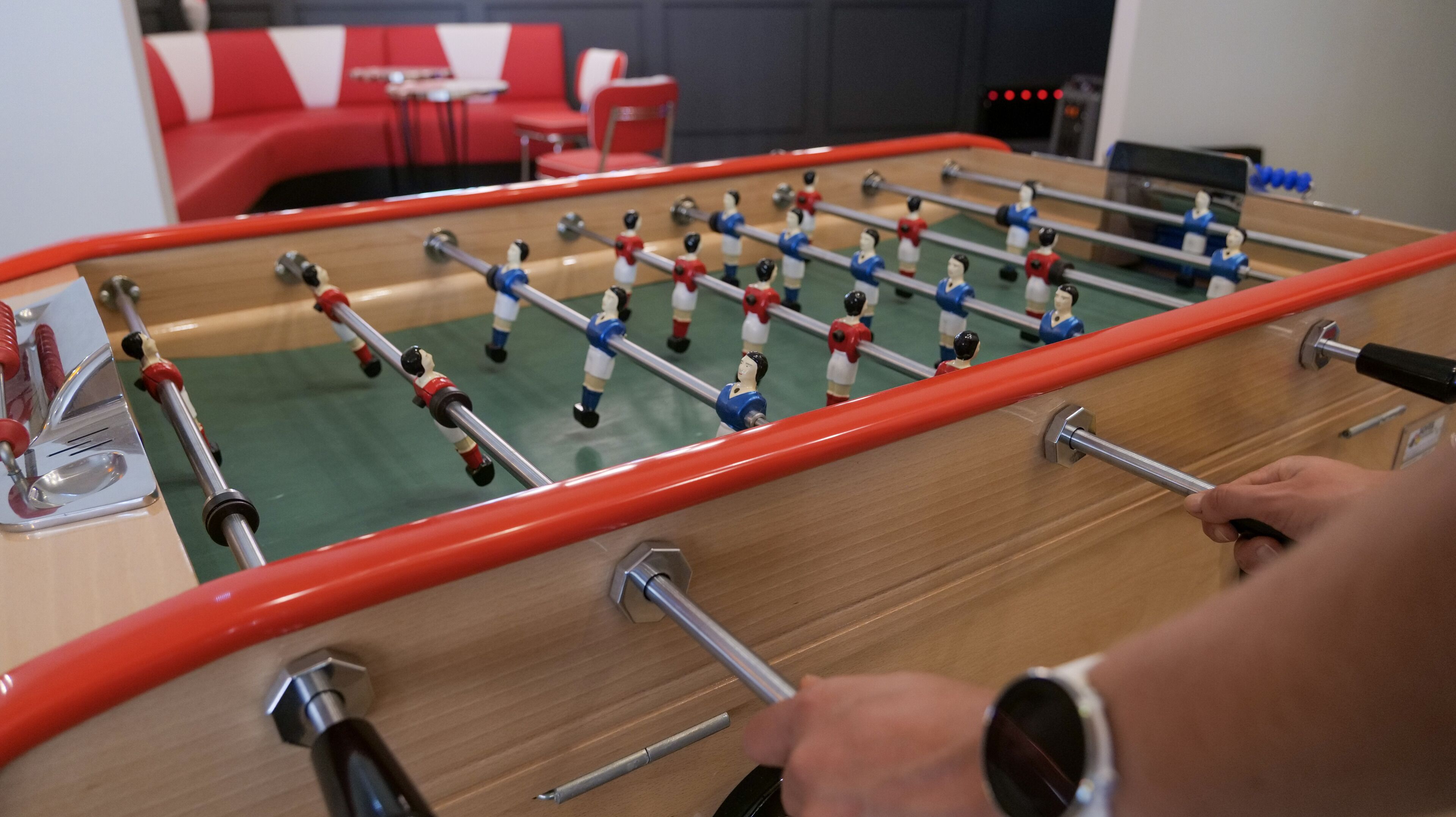 Una mesa de fútbolín con estructura roja y barras metálicas, una persona jugando, y un sofá a rayas rojas y blancas al fondo.