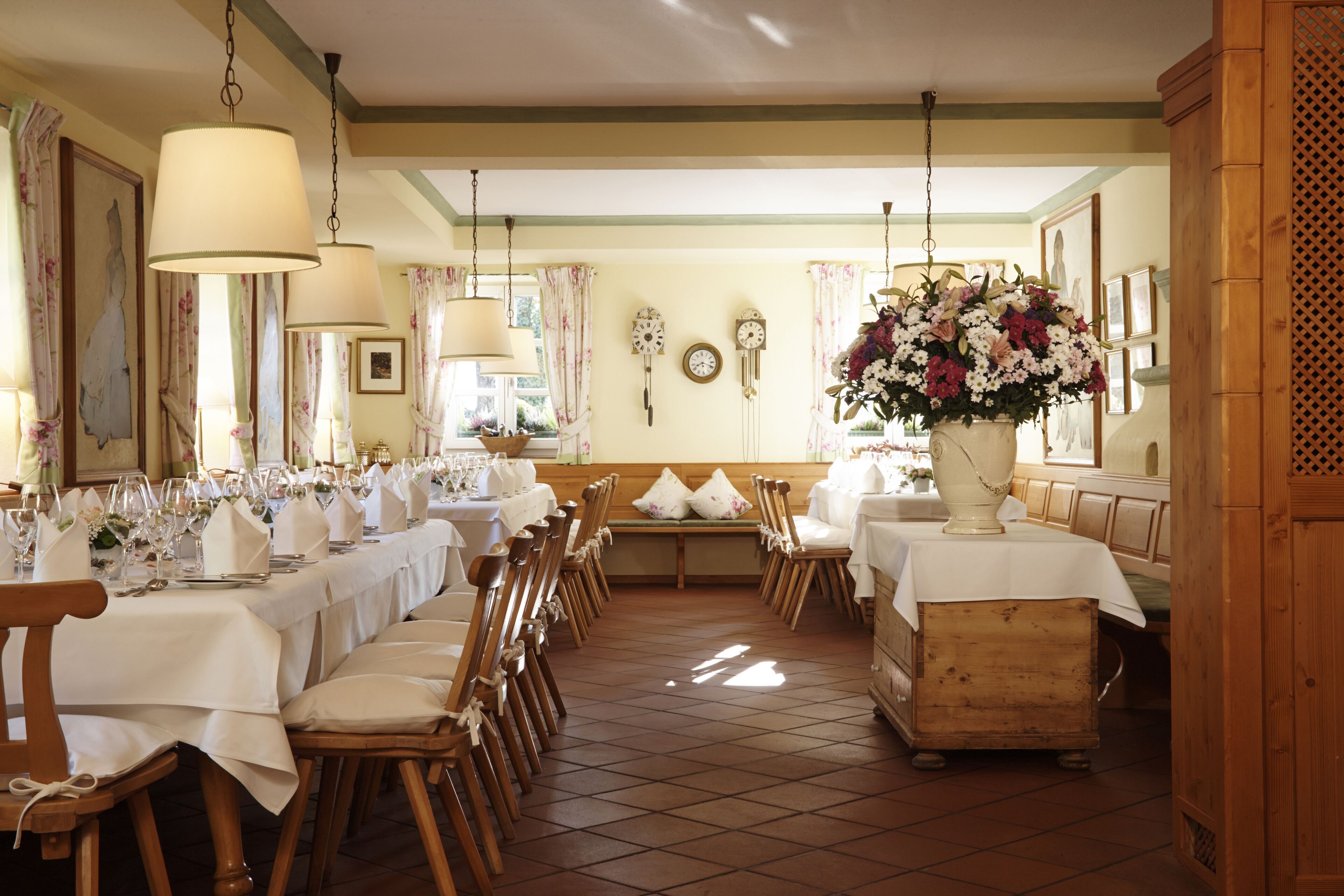 Uma sala de jantar com longas mesas de madeira preparadas para uma refeição, um grande arranjo floral e relógios de parede.