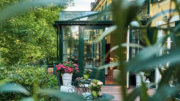 Een glazen patio met buitenmeubilair, omringd door weelderig groen en plantenpotten.